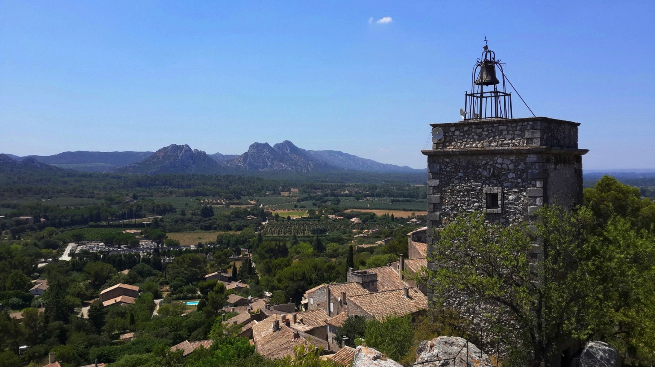 A panoramic view from a high vantage point overlooking a historic village with stone buildings, lush green valleys, and distant mountains under a clear blue sky.